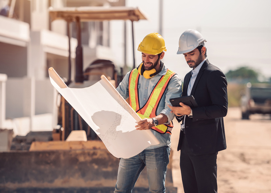 Construction collaboration: One in work attire with blueprint, the other in a suit with a phone, discussing the project.